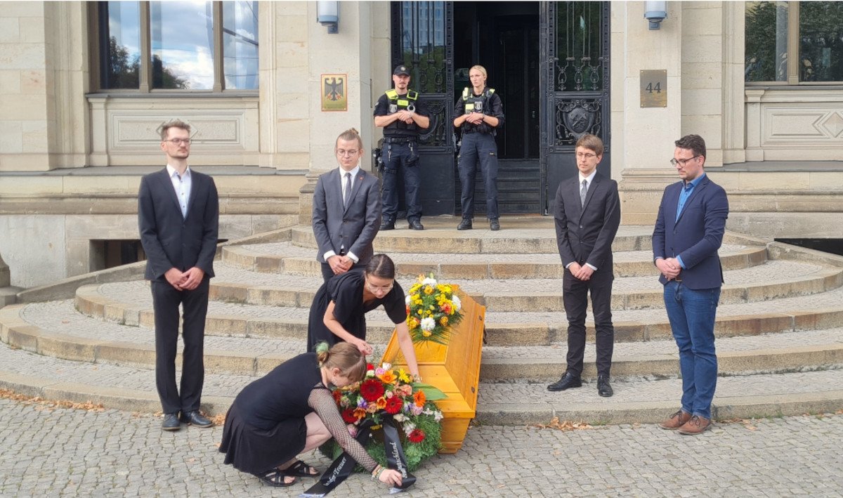 Trauernde Personen in schwarz mit einem Sarg vor einer Treppe. An den Sarg werden durch 2 der Personen Blumen nieder gelegt. 9 Euro Ticket Beisetzung