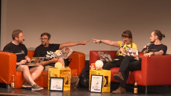 4 Personen mit 4 Laptops auf 2 roten Sofas mit viel Deko auf einer Bühne. Die beiden mittleren Personen zeigen aufeinander. Datenspuren_Live