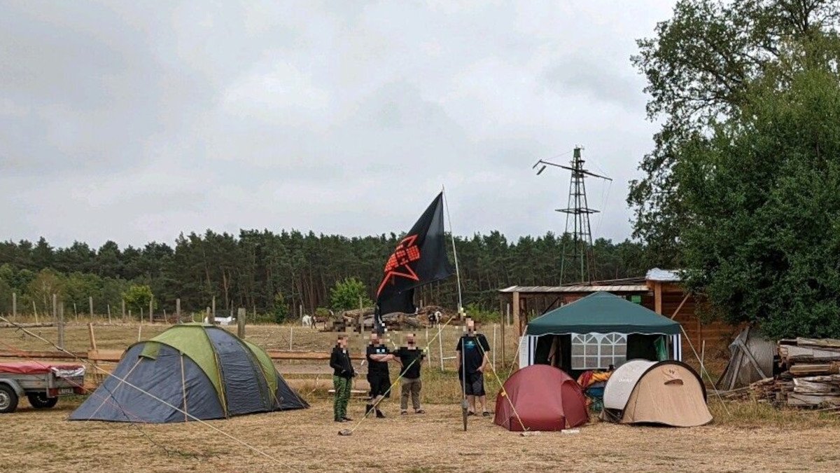 Menschen mit einer großen Fahne zwischen Zelten auf einer ausgetrockneten Wiese. Fedicamp