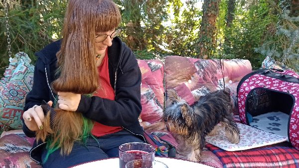 Ich, lange rotbraune Haare und große runde Brille, sitze mit einem kleinen zerzausten Hund, der mich neugierig beobachtet, auf einen Hollywood-Schaukel im Grünen. Fritzchen