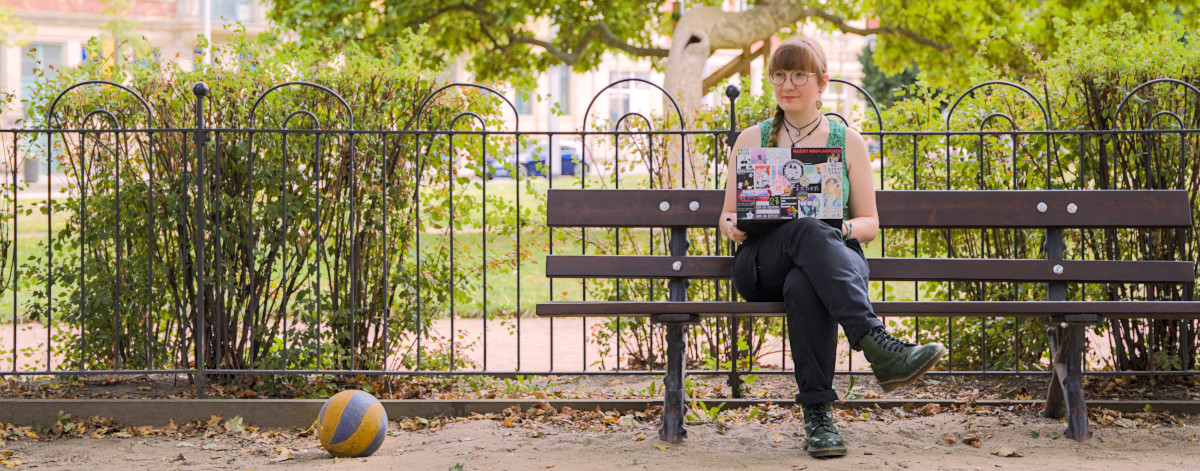 Ich, blass mit grünem Oberteil und rotbrauner Ponyfrisur mit grünen Strähnen und großer Brille, sitze mit meinem Laptop, dessen Deckel über und über mit Stickern beklebt ist, auf einer Parkbank. Links neben mir im Bild liegt ein Ball. Banner Website Ückück Stephanie Henkel